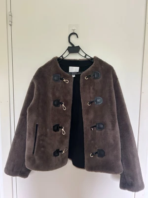 Brun pälsjacka med spännen H&M - Brun pälsjacka från H&M i mjukt fuskpälsmaterial. Jackan har rundad krage, långa ärmar och dekorativa svarta spännen med guldfärgade detaljer framtill. En ficka med dragkedja på sidan ger extra stilpoäng. Perfekt för dig som vill ha en trendig vinterlook.