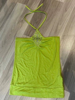 Limegrön halterneck tubtopp - Säljer en limegrön tubtopp med halterneck och dekorativa stenar framtill från ZENTE. Toppen har en triangulär detalj vid halsen och smala knytband. Står ingen storlek men skulle säga S/M. Stretchigt material. Byst: 37cm. Längd: 52cm. 