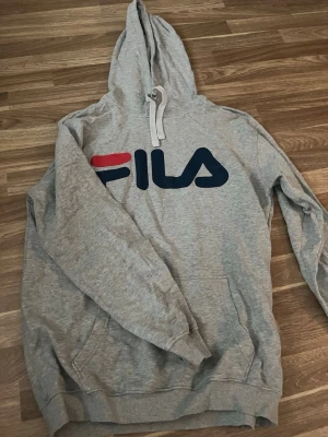Grå hoodie från FILA med logga - Säljer en grå hoodie från FILA med stor logga i blått och rött på bröstet. Tröjan har huva med vita snören och en klassisk känguruficka framtill. Perfekt för dig som gillar sportig och avslappnad stil.