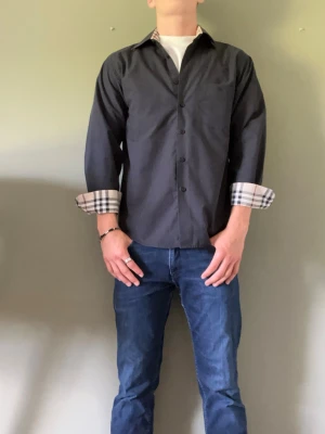Burberry skjorta - Stilren svart skjorta med klassisk krage och knappar framtill. Insidan av manschetterna har ett rutigt burberry mönster. Skjortan har lång ärm och passar perfekt till jeans eller chinos. 