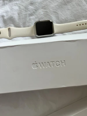 Apple Watch med flera armband - Apple Watch med silverfärgat aluminiumhölje och vit silikonrem. Medföljer extra armband i orange, blått och brunt samt laddare och adapter. Klockan har en fyrkantig skärm och stilren design, perfekt för dig som gillar smarta funktioner på handleden.