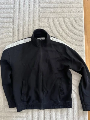 Svart Hollister zip tröja - Svart träningsjacka från Hollister med boxy fit och vita detaljer längs axlarna. Jackan har hög krage, hel dragkedja och två fickor framtill. Perfekt för sport eller chill, tillverkad i mjukt material som känns bekvämt.