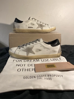 Golden Goose vita sneakers med stjärna - Säljer ett par vita Golden Goose sneakers med grå mockastjärna på sidan och svart häl. Skorna har en vintage-look med slitna detaljer och beige sula. Klassisk låg modell med snörning och lädermaterial. Perfekta för dig som gillar streetstyle och unika detaljer.
