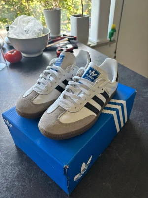 Adidas Samba OG vita och svarta - Klassiska Adidas Samba OG sneakers i vitt och svart med beige mockadetaljer på tån och gummisula. Skorna har de ikoniska tre svarta ränderna på sidan, vit ovandel i läder och blå Adidas-logga på plösen. Perfekta för en clean och tidlös streetstil.