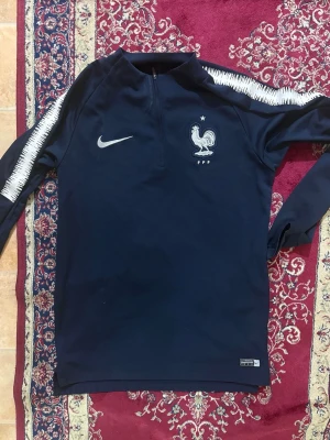 Frankrike Långärmad tröja - Mörkblå långärmad träningströja från Nike med Frankrikes landslagslogga och vit Nike-swoosh på bröstet. Tröjan har halv dragkedja och vita detaljer längs ärmarna. Perfekt för fotbollsträning eller sportiga aktiviteter.