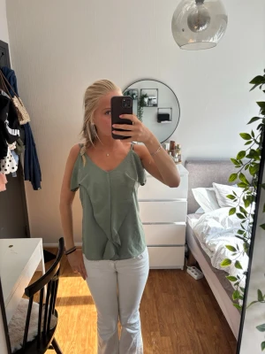 Grön linnetopp med volangdetalj - Snygg ljusgrön linnetopp med volangdetalj framtill och smala axelband. Toppen har en avslappnad passform och v-ringad halsringning. Perfekt för varma dagar och enkel att styla med jeans eller kjol.