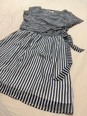 kortklänning från Tommy Hilfiger - Snygg kortklänning från Tommy Hilfiger med marinblå och vita ränder. Klänningen har korta ärmar, rund halsringning och ett knytband i midjan för en fin siluett. Perfekt för sommarens alla tillfällen.