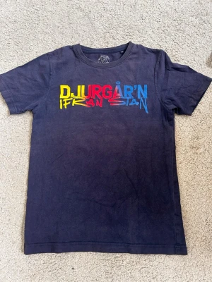 Djurgården T-shirt - Mörkblå t-shirt med färgglatt tryck på bröstet där det står 'Djurgår'n ifrån stan' i gult, rött och blått. Klassisk rund hals och korta ärmar. Perfekt till matchdagar och för dig som vill visa ditt stöd till Djurgården. 