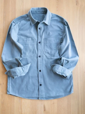 Ljusblå overshirt med fickor - Snygg ljusblå overshirt i bomull med två stora bröstfickor och svarta knappar framtill. Klassisk krage och långa ärmar med knapp vid ärmslut. Perfekt lager-på-lager-plagg för en avslappnad stil.