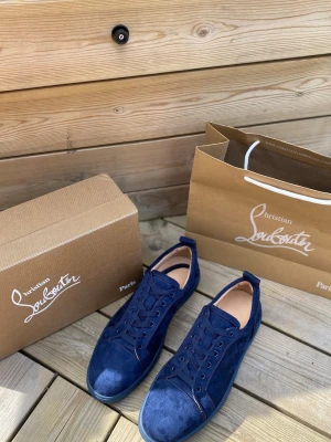 Blåa sneakers från Christian Louboutin - Mörkblå sneakers i mocka från Christian Louboutin med klassisk röd sula och ton-i-ton snörning. Skorna har en stilren och modern design med lågt skaft och diskreta detaljer. Perfekta för dig som vill ha något exklusivt och snyggt till din outfit.