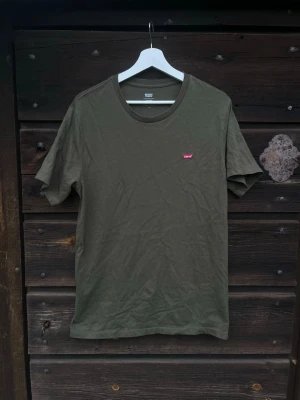 Olivgrön t-shirt från Levi's - Säljer en enkel och stilren olivgrön t-shirt från Levi's med klassisk passform och liten röd logotyp på bröstet. T-shirten har rund halsringning och korta ärmar, perfekt till jeans eller shorts.