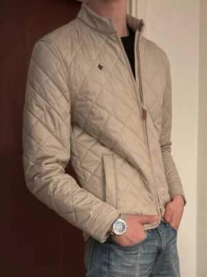 Morris Jacka  - Säljer en mycket fin quiltad jacka från Morris i en elegant beige nyans. Jackan har en lätt och smidig dunfyllning som ger lagom värme utan att kännas tung, vilket gör den perfekt för svala sommarkvällar, tidiga höstdagar eller som ett stilrent lager under större delen av året.  Den klassiska quiltade designen tillsammans med Morris diskreta logotyp ger ett tidlöst och välklätt intryck som fungerar lika bra till jeans och sneakers som till en mer uppklädd outfit.