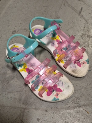 Sandaler - Säljer ett par färgglada sandaler från Gios Eppo med transparenta rosa remmar och turkosa detaljer. Sulan har ett lekfullt fjärilsmönster i flera färger. Tillverkade i plastmaterial och perfekta för sommarens alla äventyr.