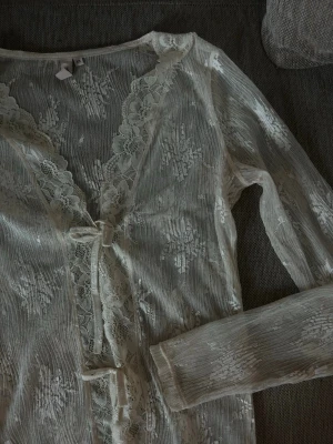 Vit spetsblus med knyt XS - Säljer en superfin vit spetsblus i transparent mesh med långa ärmar och vacker blommig spetskant längs v-ringningen. Blusen har knyt framtill och ger en romantisk vibe. 