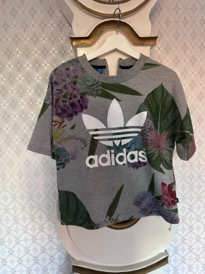 Grå blommig Adidas t-shirt - Säljer en grå t-shirt från Adidas med stort vitt logotryck på bröstet och färgglatt blommönster i grönt, lila och rött. T-shirten har korta ärmar och en croppad passform. Perfekt för dig som vill sticka ut lite extra!