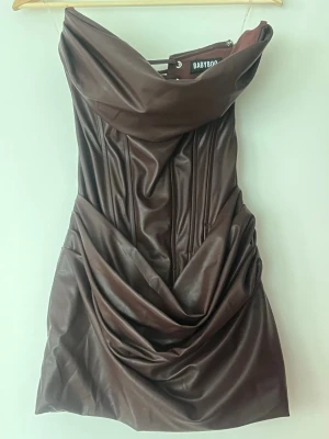 Brun axelbandslös klänning Babyboo - Säljer en brun axelbandslös kort klänning från Babyboo i ett glansigt syntetmaterial med draperad kjol och markerad midja. Klänningen har en snygg omlott-detalj framtill och en strukturerad, figurnära passform som ger en riktigt trendig look.