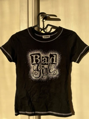 Svart t-shirt med Bad Girl-tryck - Svart t-shirt med vit kontrastsöm och ett stort 'Bad Girl'-tryck i glitter framtill. T-shirten har korta ärmar och rund halsringning. 