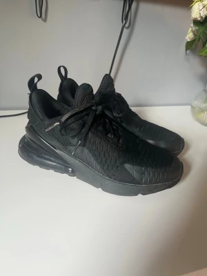 Nike Air Max 270 helsvarta sneakers - Säljer ett par helsvarta Nike Air Max 270 sneakers med mesh-ovandel och synlig Air-enhet i hälen. Skorna har en sportig look med snörning och dragflik bak. Perfekta för dig som gillar stilrena och bekväma sneakers.