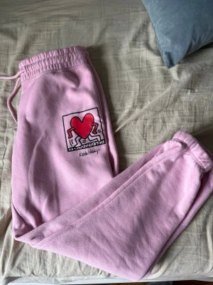 Rosa mjukisbyxor Keith Haring  - Säljer ett par ljusrosa mjukisbyxor med elastisk midja och dragsko. På ena benet finns ett ikoniskt Keith Haring-tryck med en röd hjärtfigur och svarta konturer. Byxorna är i mjukt bomullsmaterial och har avslappnad passform. 