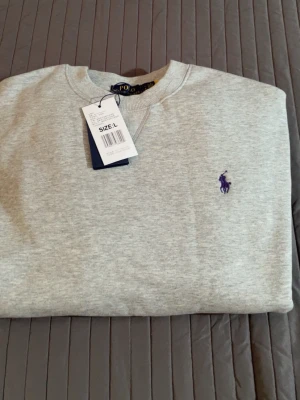 Grå sweatshirt från Polo Ralph Lauren - Snygg grå sweatshirt från Polo Ralph Lauren med klassisk rund hals och lila broderad logga på bröstet. Tröjan har ribbade muddar vid ärmslut och nederkant, perfekt för en avslappnad och stilren look. Materialet är mjukt och bekvämt.