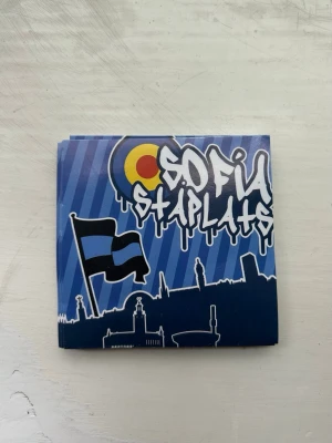 Sofia Ståplats klistermärke - Klistermärke med texten 'Sofia Ståplats' i graffiti-stil, blå bakgrund med siluett av stad och svart flagga med blått kors. Perfekt för att dekorera laptop, mobil eller anteckningsbok.