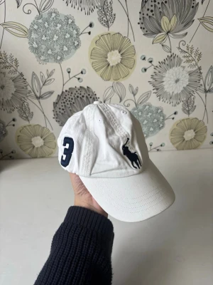 Vit keps från Polo Ralph Lauren - Säljer en vit keps från Polo Ralph Lauren med den klassiska broderade loggan i marinblått framtill och en stor siffra 3 på sidan. Kepsen har böjd skärm och justerbar rem bak. 