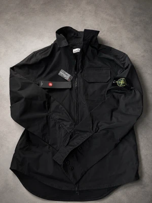 Svart overshirt från Stone Island - Snygg svart overshirt från Stone Island med dragkedja framtill och klassisk patch på vänster ärm. Overshirten har en bröstficka med lock och är gjord i ett slitstarkt material som ger en clean och modern look. Perfekt lager på lager-plagg för streetwear-stilen.