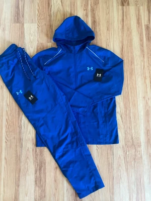 Blått träningsset från Under Armour - Snyggt blått träningsset från Under Armour med matchande hoodie och byxor. Setet har dragkedja på tröjan, huva och vita detaljer på axlarna. Tillverkat i lätt och ventilerande funktionsmaterial, perfekt för träning eller chill.