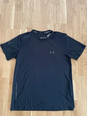 Svart träningströja från Under Armour - Svart kortärmad träningströja från Under Armour med diskret logga på bröstet och tryck på ärm och nederkant. Tillverkad i ett lätt och snabbtorkande funktionsmaterial som passar perfekt för träning och sport. Den passar även M, L, XL beroende på hur man vill den ska sitta.