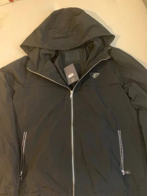 Svart vindjacka - Snygg svart vindjacka  med huva och dragkedja framtill. Jackan har två fickor med silverfärgade dragkedjor och den klassiska har en logga på bröstet. Tillverkad i lätt och tåligt material, perfekt för blåsiga dagar. Den är M men passar S lite mer. Byten går och priset kan diskuteras.