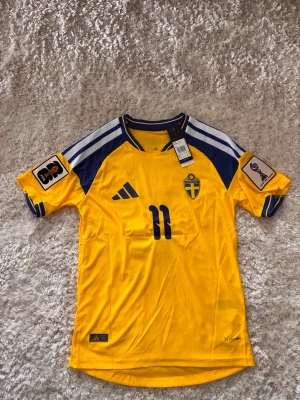 Svensk landslagströja Adidas gul - Gul svensk landslagströja från Adidas med blå detaljer och nummer 11 på bröstet. Tröjan har officiella FIFA-märken på ärmarna, blåa ränder på axlarna och det svenska landslagets emblem på bröstet. Tillverkad i lätt och ventilerande material.