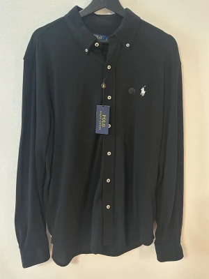 Svart skjorta från Polo Ralph Lauren - Klassisk svart skjorta från Polo Ralph Lauren med vit broderad logga på bröstet och vita knappar. Skjortan har lång ärm och normal passform, perfekt till en stilren look. Tillverkad i mjuk bomull för skön känsla. Helt oanvänd prislappen är kvar.