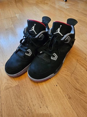Air Jordan 4 Retro Black/Red - Säljer ett par Air Jordan 4 Retro sneakers i svart mocka med grå och röda detaljer. Skorna har den klassiska Jumpman-loggan på plösen och meshpaneler på sidorna. Sulan är vit och röd med synlig Air-enhet. Perfekt för dig som gillar streetwear och ikoniska modeller. Kan prata om priset
