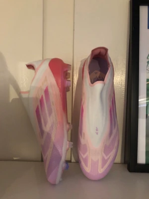 Adidas rosa och vita fotbollsskor - Säljer ett par Adidas fotbollsskor med en snygg design i rosa och vitt. Skorna har en strömlinjeformad siluett, utan snörning, och är gjorda i syntetmaterial med plastdobbar för bra grepp på planen. Perfekta för dig som vill sticka ut på matchen.