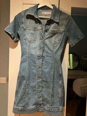 Kort jeansklänning från Zara - Snygg kort jeansklänning från Zara med knappar framtill, två bröstfickor och klassisk skjortkrage. Klänningen har korta ärmar och markerad midja för en feminin siluett. Perfekt för en trendig och avslappnad look.