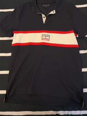 Marinblå piké från Tommy Hilfiger - Snygg marinblå pikétröja från Tommy Hilfiger med vit och röd rand över bröstet och logga i mitten. Klassisk krage med knappar och korta ärmar. Perfekt för en avslappnad men stilren look.