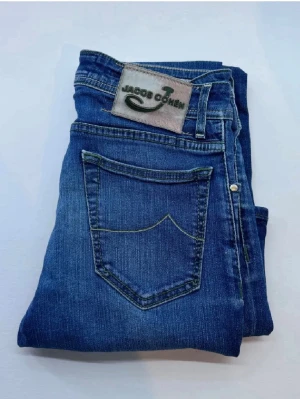 Blå jeans från Jacob Cohen - Snygga blå jeans från Jacob Cohen med klassisk femficksdesign och broderad logga på bakfickan. Jeansen har knappgylf med silverfärgade knappar och detaljerad etikett bak i midjan. Tillverkade i mjukt denimtyg för en bekväm känsla. 