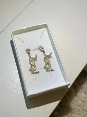 Glittriga YSL örhängen med stenar - Säljer ett par ikoniska YSL örhängen i guldton med gnistrande stenar. Hängande modell med YSL-logga täckt av små glittrande stenar som ger en lyxig känsla. Perfekta för dig som vill sticka ut och addera lite extra bling till din look.OBS! Ej rostfria. Kan rosta/oxidera med tiden. Genomskinligt nagellack rekommenderas som skydd.