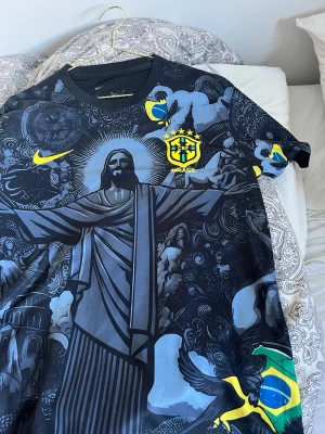 Brasil Nike fotbollströja svart/grå - Snygg svart och grå fotbollströja från Nike med Brasilien-tema. Tröjan har ett stort tryck av Kristusstatyn, brasilianska flaggan och detaljer i gult. CBF-emblemet är broderat på bröstet. Perfekt för dig som älskar fotboll och vill sticka ut på planen.