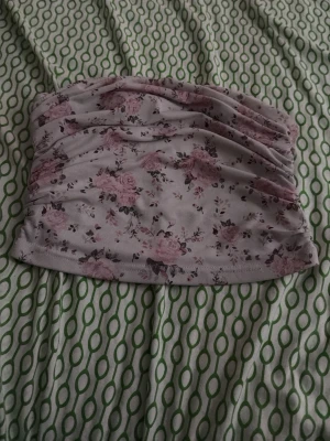 Ljus blommig bandeau tubtopp - Supersöt bandeau tubtopp i ljusrosa och vitt med blommigt mönster. Toppen är ärmlös och har rynkade detaljer framtill för en snygg passform. Perfekt för varma dagar och enkel att styla med jeans eller kjol.
