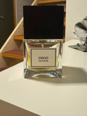 D600 Eau de Parfum från Carner - D600 Eau de Parfum från Carner Barcelona i en stilren, fyrkantig glasflaska med svart fyrkantigt lock. Flaskan har en transparent design som visar den gyllengula vätskan inuti och en enkel vit etikett framtill. Doften är exklusiv och flaskan känns lyxig och modern.