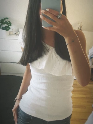 Vit offshoulder topp med volang - Säljer en vit offshoulder topp med volangdetalj över bysten. Toppen är ribbad och har en tight passform som framhäver figuren. Perfekt för dig som vill ha en stilren men ändå trendig look. En en lite roligare basic vit topp som klär upp outfitsen. Storleken är 164 men sitter som XS.