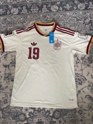 Vit Spanien fotbollströja Adidas - Säljer en vit Spanien fotbollströja från Adidas med vinröda detaljer, nummer 19 och Lamine Yamal på ryggen. Tröjan har gula och vinröda ränder på ärmslut och krage, samt broderad landslagssköld och Adidas-logga på bröstet. Perfekt för match eller träning.