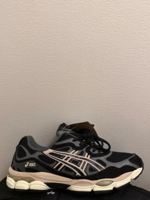 Svarta Asics sneakers med beige detaljer - Snygga svarta sneakers från Asics med beige och grå detaljer. Skorna har en chunky sula i vitt och beige, meshpaneler för bra ventilation och klassiska Asics-loggan på sidan. Perfekta för dig som gillar sportig och trendig stil.