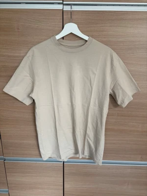 Beige basic t-shirt i bomull - Enkel och stilren beige t-shirt med rund hals och korta ärmar. Tillverkad i mjuk bomull för en skön känsla. Perfekt att matcha med jeans eller shorts för en clean look.