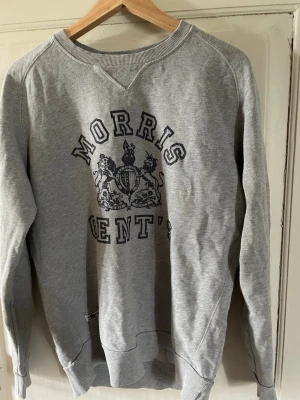 Grå sweatshirt från Morris - Klassisk grå sweatshirt från Morris med tryckt logga och emblem på bröstet. Tröjan har rund halsringning, ribbade muddar vid ärmslut och nederkant samt en avslappnad passform. Perfekt för en casual look.