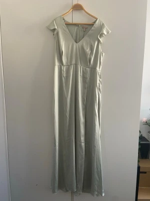Ljusgrön långklänning  - Elegant ljusgrön långklänning med v-ringad hals och korta volangärmar. Klänningen har en mjuk, något glansig yta och ett flytande fall som ger en romantisk känsla. Perfekt för sommarens alla tillfällen.