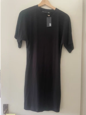 Svart t-shirtklänning från 157 - En enkel och stilren svart t-shirtklänning från 157 med rund halsringning och korta ärmar. Klänningen har en kropps passform och är gjord i mjukt bomullsmaterial som känns skönt mot huden. Perfekt för dig som gillar minimalistisk stil.