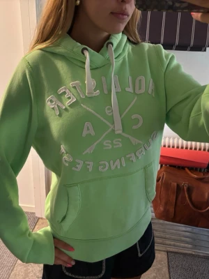Grön hoodie från Hollister - Snygg ljusgrön hoodie från Hollister med vit broderad text och logga på bröstet. Hoodien har huva med vita dragsnören och en stor magficka. Perfekt för en avslappnad och sportig stil. Mjuk bomullsblandning som känns skön mot huden.