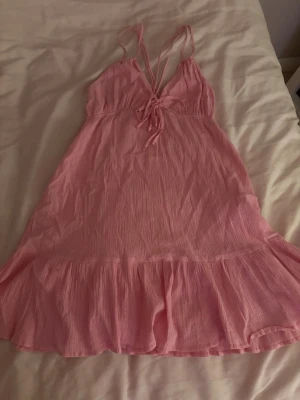 Rosa kort sommarklänning med volang - Söt rosa kortklänning med tunna axelband som korsas i ryggen och knytning framtill. Klänningen har volangkant nertill och är gjord i ett lätt, luftigt tyg som passar perfekt för varma dagar.
